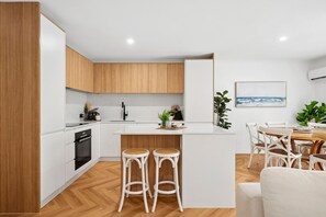 Eine moderne Küche mit weißen Schränken, einer zentralen Kochinsel mit Hockern, einem Essbereich mit Holztisch und Stühlen sowie einem Gemälde an der Wand.