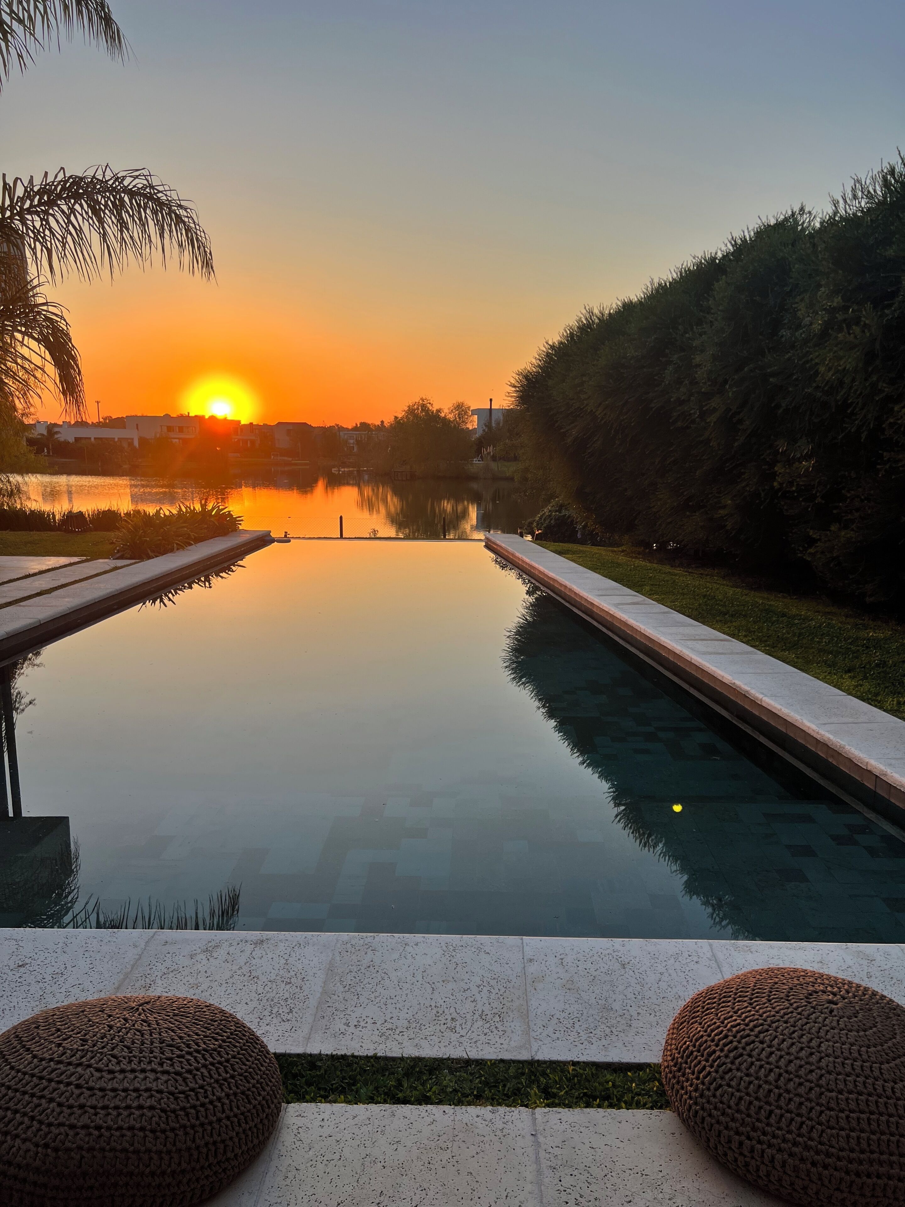 A serene outdoor pool area with a sunset view, surrounded by greenery and a clear sky.