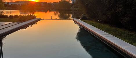 A serene outdoor pool area with a sunset view, surrounded by greenery and a clear sky.
