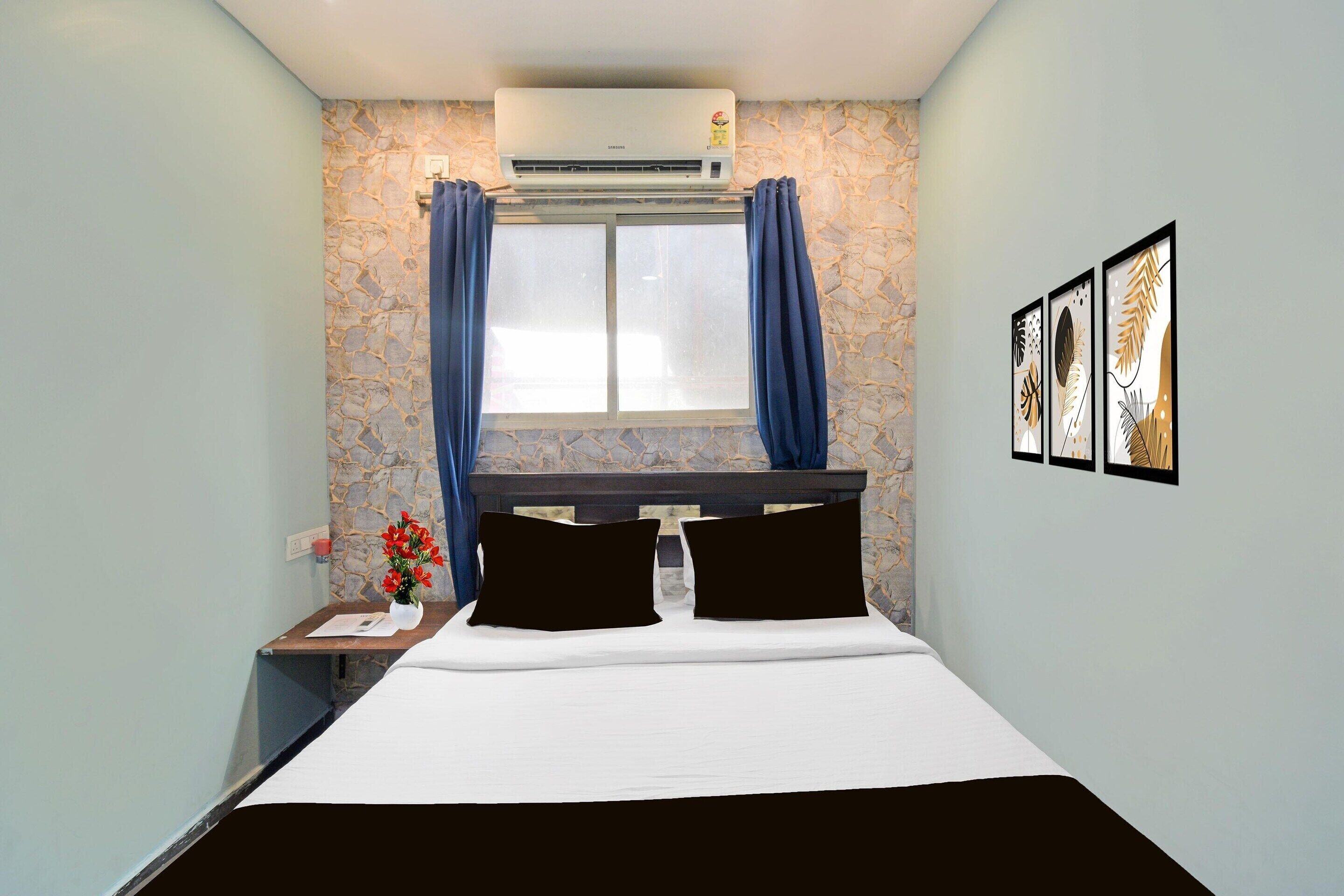 Ein Hotelzimmer mit einem Bett, einer Klimaanlage, einem Fenster mit Vorhängen, einem Nachttisch mit Blumenvase und gerahmten Kunstwerken an der Wand.