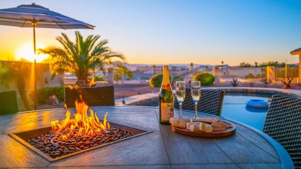 Een tafel met een vuurplaats, een fles en glazen, met uitzicht op een zwembad bij zonsondergang.