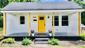 A white single-story house with a yellow door, number 1200, and a porch with potted plants.
