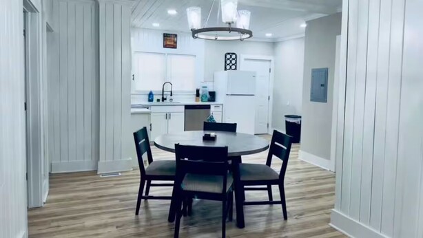 A dining area with a round table and chairs, a kitchen in the background, and a chandelier.