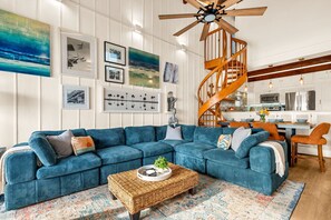 A living room with a blue sectional sofa, a wooden spiral staircase, and a kitchen in the background. - Oceanfront Condo | Rooftop Terrace | Arcades | (Indian Shores)