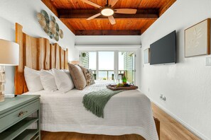 Quarto com cama, cabeceira de madeira, ventilador de teto, TV e vista para o exterior.