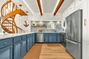 A modern kitchen with blue cabinets, a stainless steel refrigerator, wooden ceiling beams, and a spiral staircase. - Oceanfront Condo | Rooftop Terrace | Arcades | (Indian Shores)