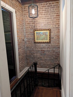 A staircase with a wooden handrail, a black metal railing, a brick wall, and a hanging light fixture.