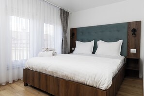 A modern hotel room with a wooden bed frame, white bedding, and a view of the city through sheer curtains. - Kulla e Sahatit in Old Bazaar (Gjakova)