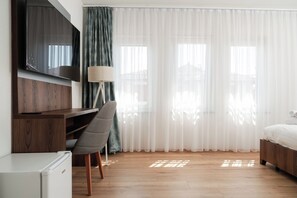 Ein Hotelzimmer mit einem Bett, einem Schreibtisch mit Stuhl, einem Fernseher und einem Fenster mit durchsichtigen Vorhängen.
