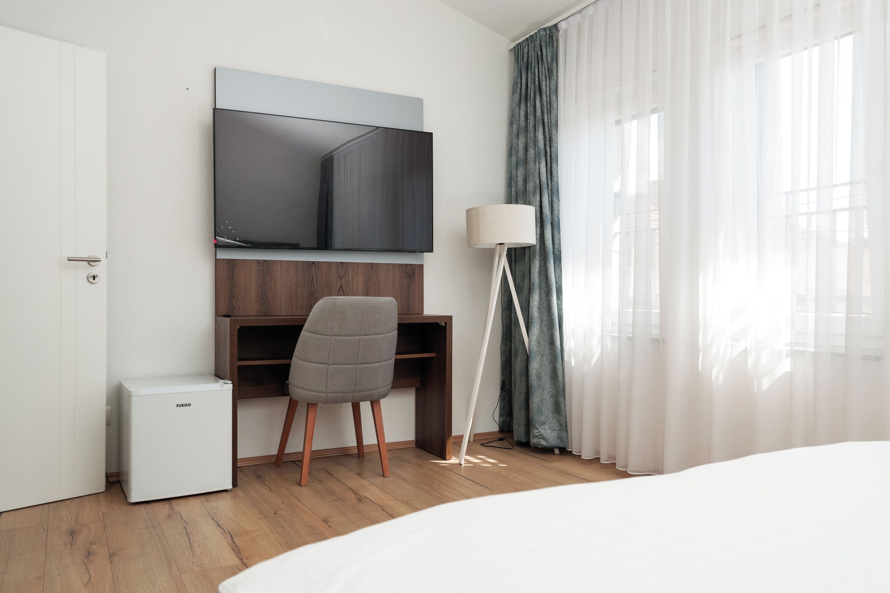 Une chambre d’hôtel comprenant un lit, un bureau avec une chaise, un téléviseur fixé au mur, un petit réfrigérateur et une lampe sur pied.