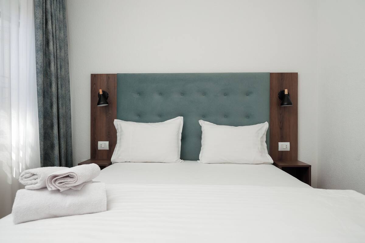 A hotel room with a bed, two bedside tables, a headboard, and a window with curtains.