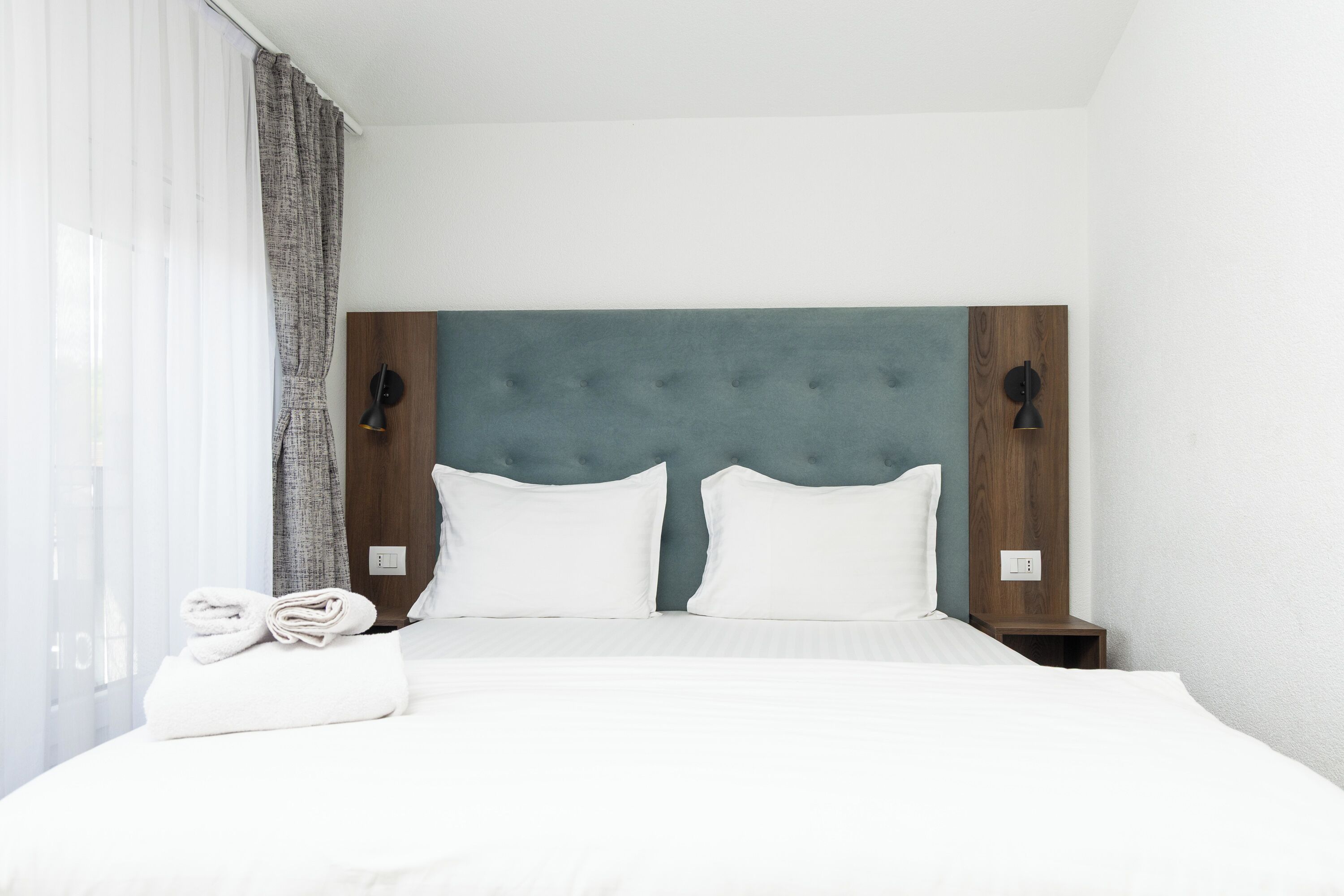A neatly made bed with white linens, a grey upholstered headboard, and two bedside lamps.