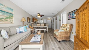 A living room with a sofa, coffee table, wicker chair, and a dining area with a table and chairs. - Steps to the Sand: Romar Beach 106 (Orange Beach)