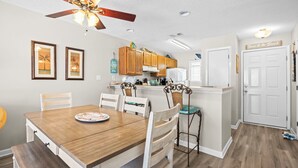 A dining area with a wooden table and chairs, a kitchen with wooden cabinets, a ceiling fan, and a door with a sign. - Steps to the Sand: Romar Beach 106 (Orange Beach)