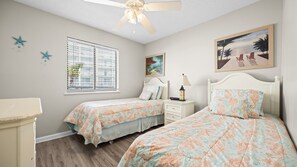 A bedroom with two beds, a ceiling fan, a window with blinds, a bedside table with a lamp, and a painting on the wall. - Steps to the Sand: Romar Beach 106 (Orange Beach)