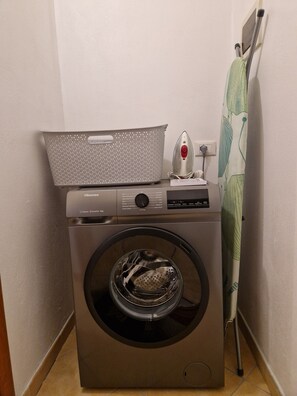 Máquina de lavar com carga frontal, um cesto no topo, um centro de engomar fixado na parede e uma tomada.