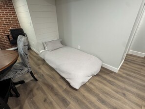 Une petite chambre avec un lit simple, un plancher en bois, un bureau avec une chaise et un mur en briques.