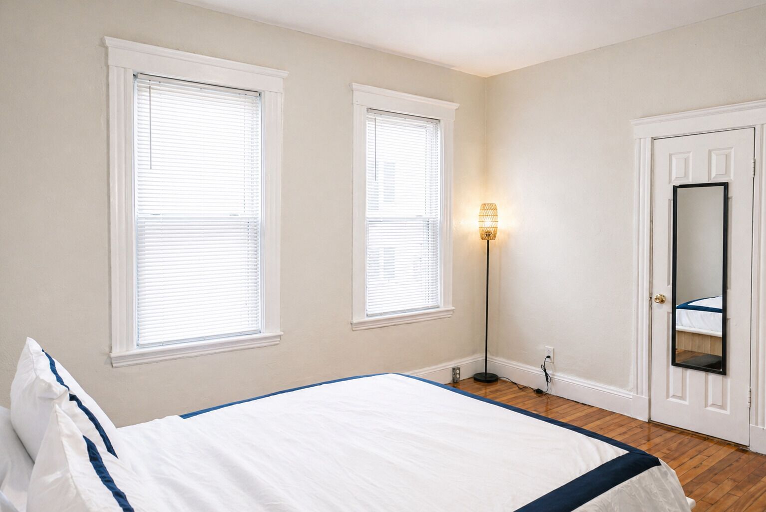 Quarto com cama, duas janelas, abajur, porta com espelho e parede branca.