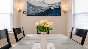 A dining table with a vase of white tulips, two chairs, and a framed painting of a seascape.