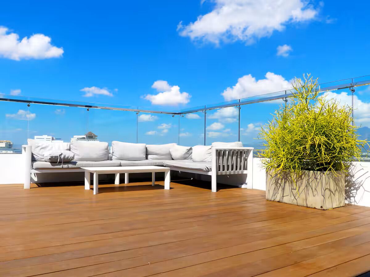 A wooden deck with a white sofa and a small table, a potted plant, and a clear blue sky.
