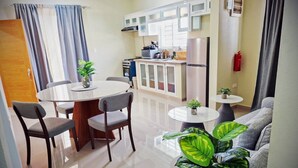 A modern kitchen with white cabinets, a stainless steel refrigerator, and a gas stove. There is a dining area with a round table and chairs, and a living area with a sofa and a large plant. - MB apartment  (Azua)