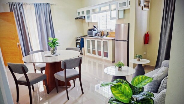 A modern kitchen with white cabinets, a stainless steel refrigerator, and a gas stove. There is a dining area with a round table and chairs, and a living area with a sofa and a large plant. - MB apartment  (Azua)