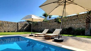 Uma piscina com espreguiçadeiras e guarda-sóis, cercada por um muro de pedra e vegetação.