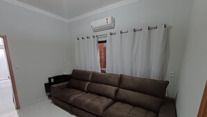 A room with a brown sofa, white curtains, and an air conditioning unit. - Charming house near the state hospital, bakery, supermarket, and pharmacy. (franca)