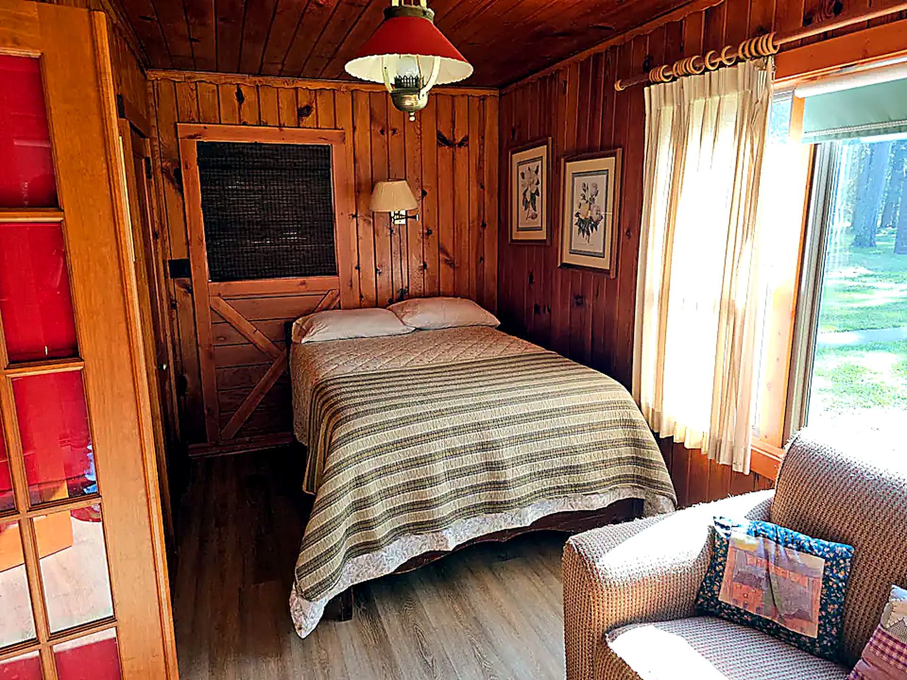 A wooden cabin room with a bed, a chair, a window with curtains, and a hanging light fixture.
