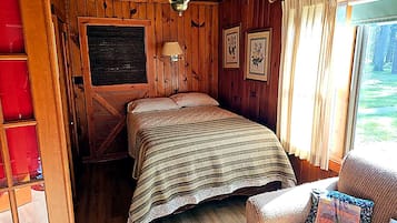 A wooden cabin room with a bed, a chair, a window with curtains, and a hanging light fixture.