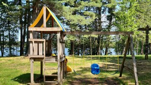 Een houten speelconstructie met een glijbaan en schommels, gelegen in een grasrijke omgeving met bomen en een meer op de achtergrond.