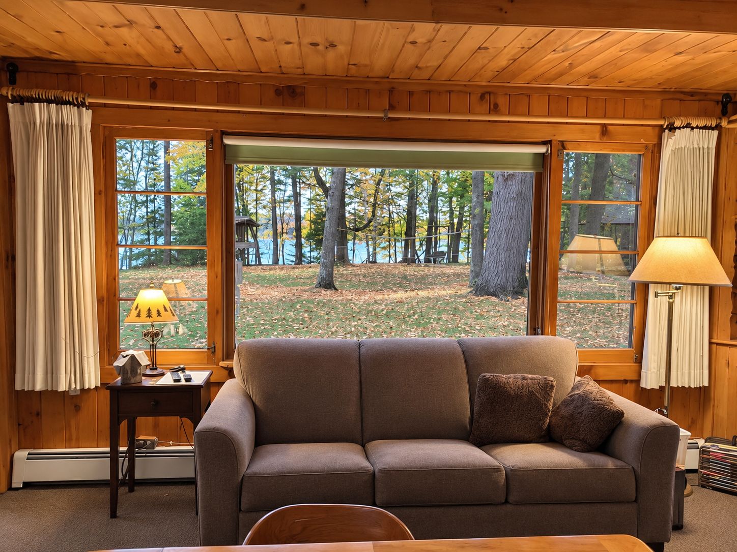 Ein gemütliches Zimmer mit Holzbalkendecke, einem braunen Sofa mit Kissen, einem kleinen Tisch mit einer Lampe und großen Fenstern mit Blick auf Bäume und einen Garten.