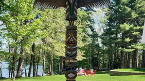 A totem pole with various faces and wings, set in a grassy area with trees and a lake in the background.