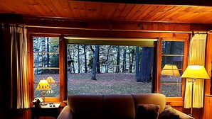 A cozy cabin interior with a wooden ceiling, a sofa, a table with a lamp, and a view of a forest through large windows.