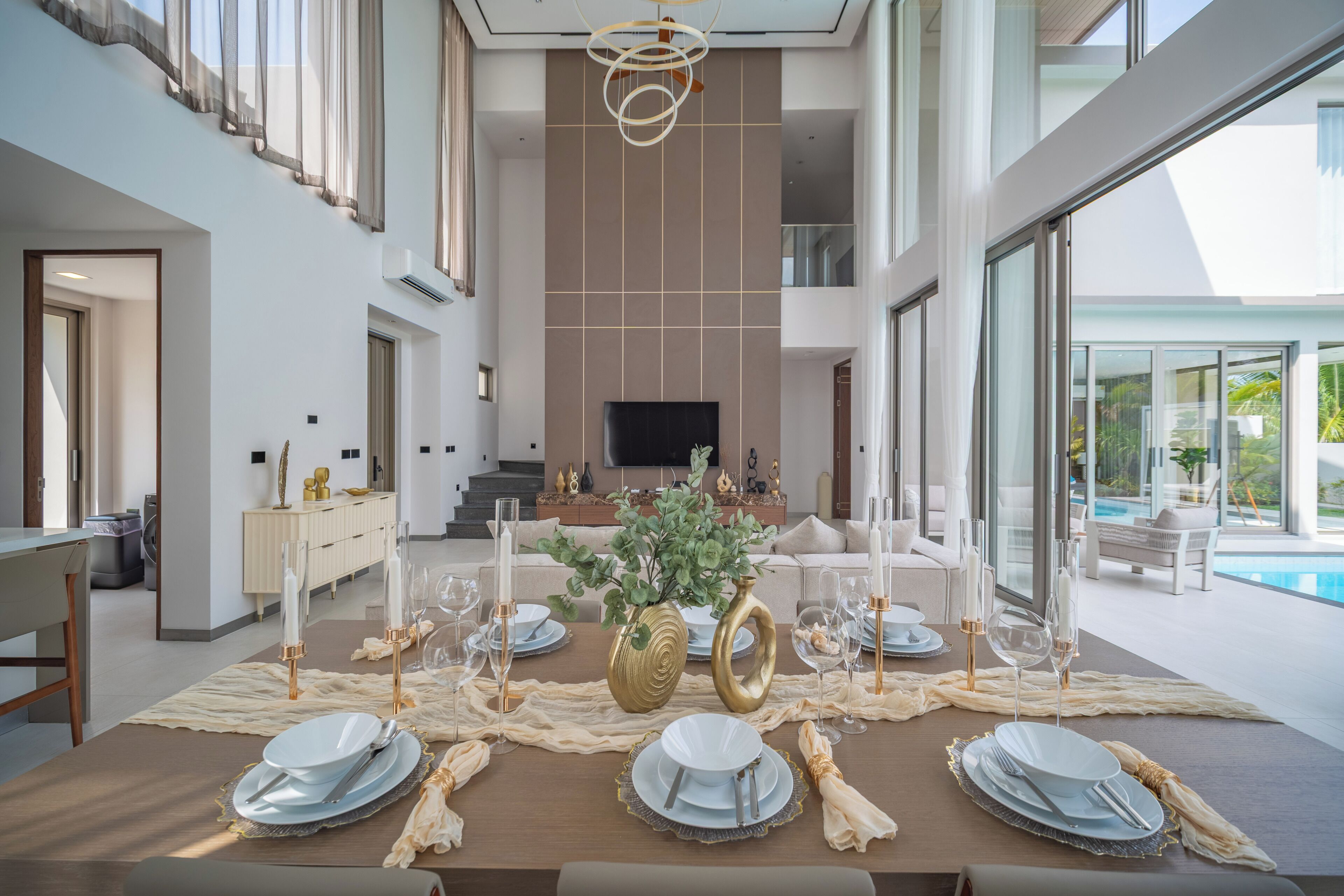Un comedor con una mesa larga dispuesta para seis personas, un televisor al fondo y una piscina visible a través de amplias ventanas.