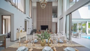 A dining area with a long table set for six, a TV in the background, and a pool visible through large windows. - GRAND VIEW Residences Villa with Pool (Choeng Thale)