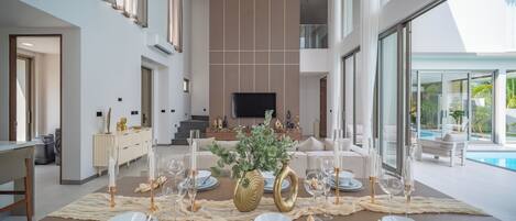 Un comedor con una mesa larga dispuesta para seis personas, un televisor al fondo y una piscina visible a través de amplias ventanas.