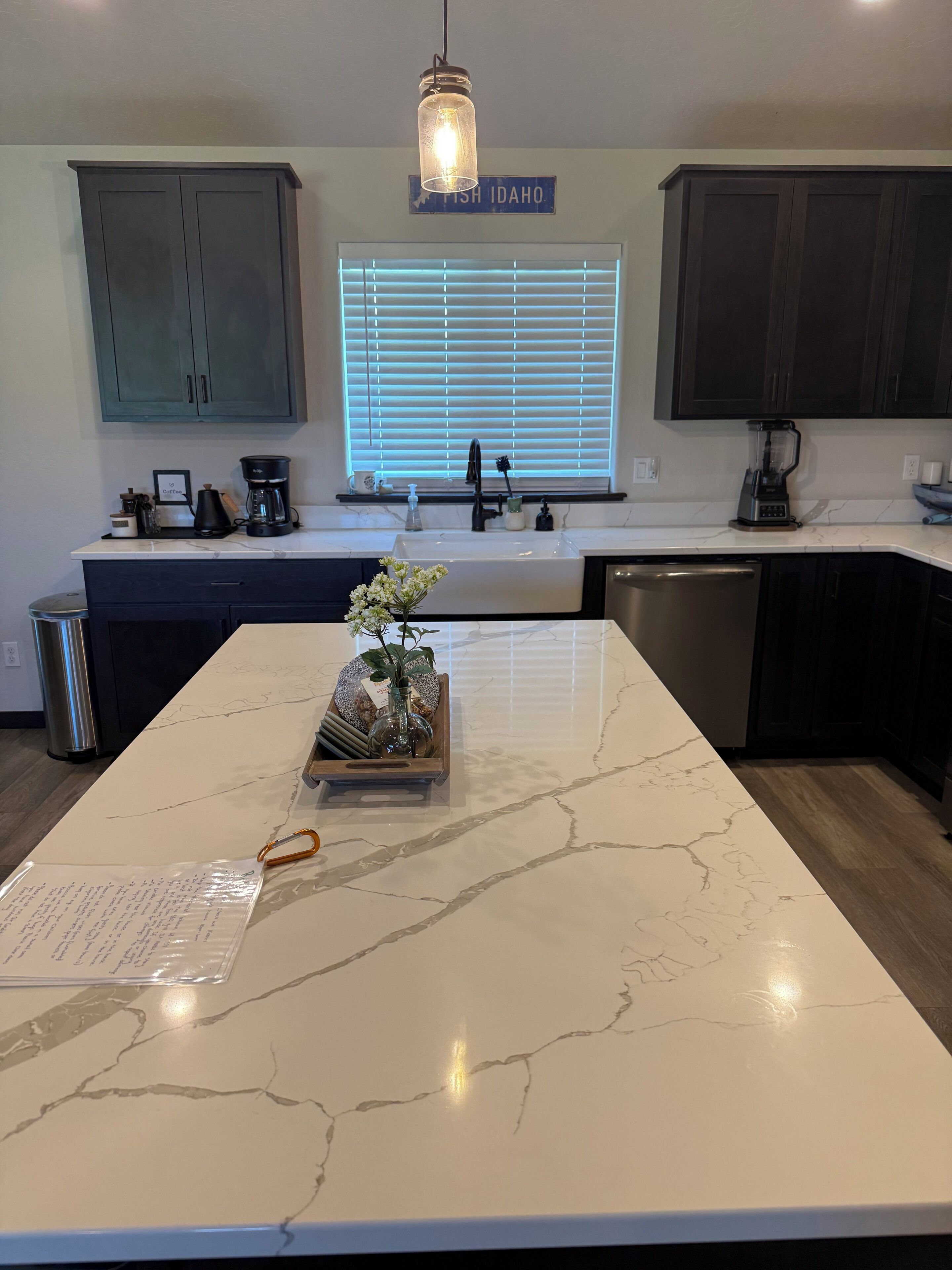 A kitchen with a marble island, dark cabinets, a window with blinds, and a hanging light fixture.