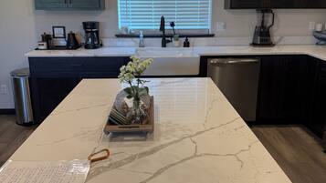 A kitchen with a marble island, dark cabinets, a window with blinds, and a hanging light fixture.
