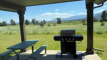 A covered outdoor area with a grill, picnic table, and bench, overlooking a grassy field and mountains.