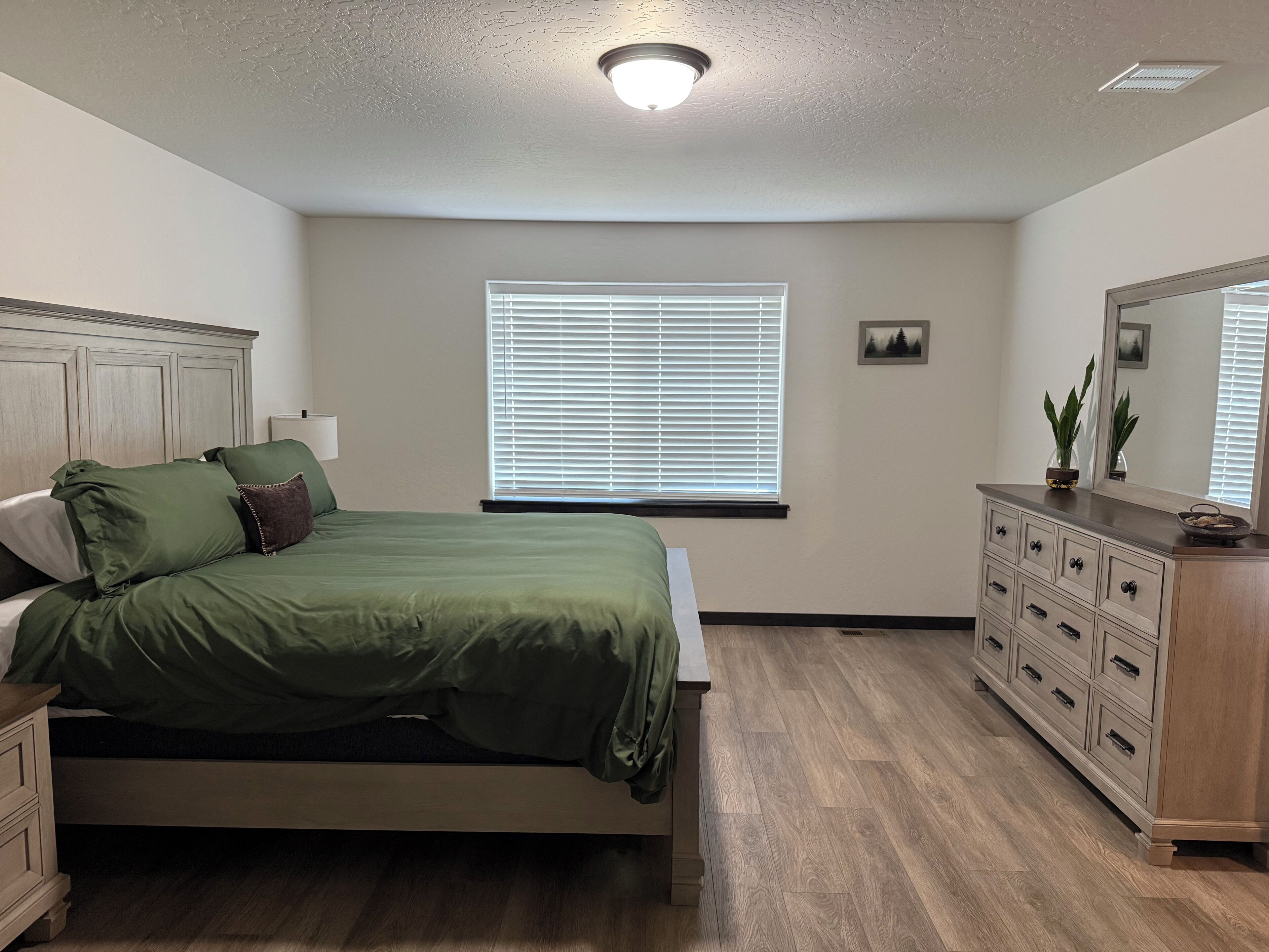 A bedroom with a bed, dresser, window with blinds, and a ceiling light.