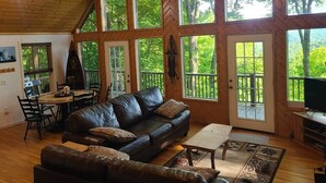 A spacious living room with a large leather sofa, a dining table, and multiple windows offering views of a forested area. - Fall colors,mountain views, family-friendly cabin, hot tub, kayaks, on two acres (Topton)