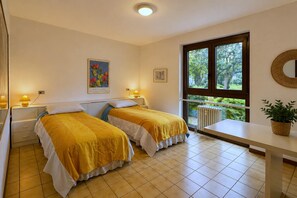 A room with two beds, a window with a view of greenery, a bedside table with a lamp, and a wall-mounted mirror. - Spacious Apartment in Luino for 5 (Luino)