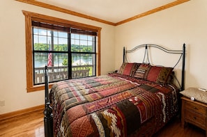 Ein Schlafzimmer mit großem Fenster, einem Bett mit gemusterten Bettbezügen, einem Nachttisch und Holzboden.