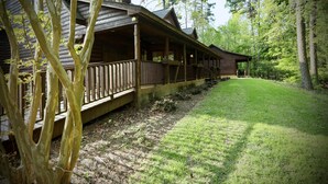 Une cabane en rondins avec un porche, entourée d’arbres et bordée d’une pelouse.