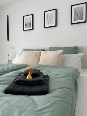 A neatly made bed with green bedding, two pillows, and a folded towel with a flower.