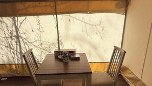 Ein Zelt mit Tisch und Stühlen, ein kleiner Tisch mit einer Kiste und einer Tasse sowie ein Fenster mit Blick auf kahle Bäume.