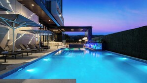 Piscina com espreguiçadeiras e guarda-sóis, área de bar e vista da cidade ao entardecer.