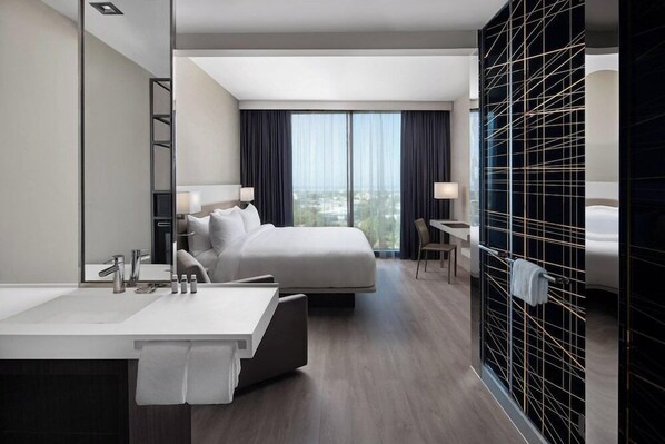 A modern hotel room with a large bed, a separate sitting area, a large window with curtains, a bathroom with a mirror and sink, and a black tiled wall. - AC Hotel Kingston Jamaica (Kingston)