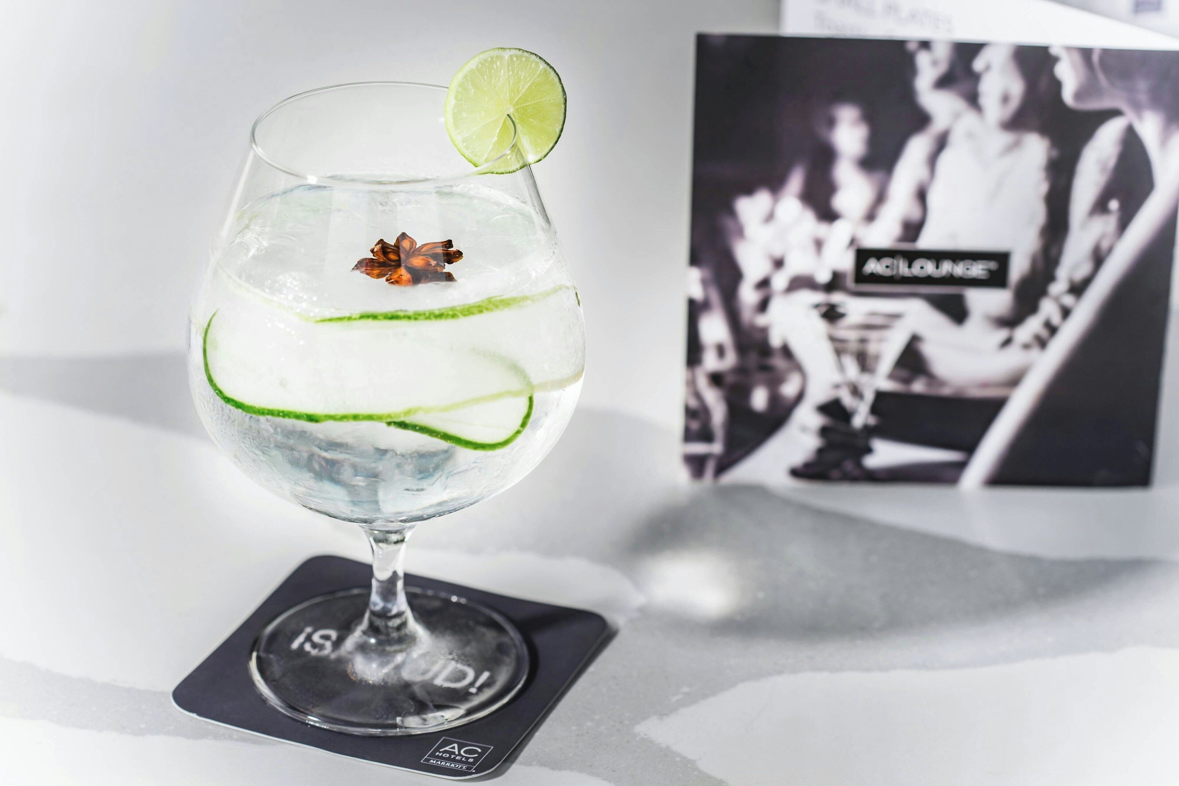 A cocktail glass with a lime garnish, star anise, and green rind slices on a coaster with 'AC LOUNGE' branding.